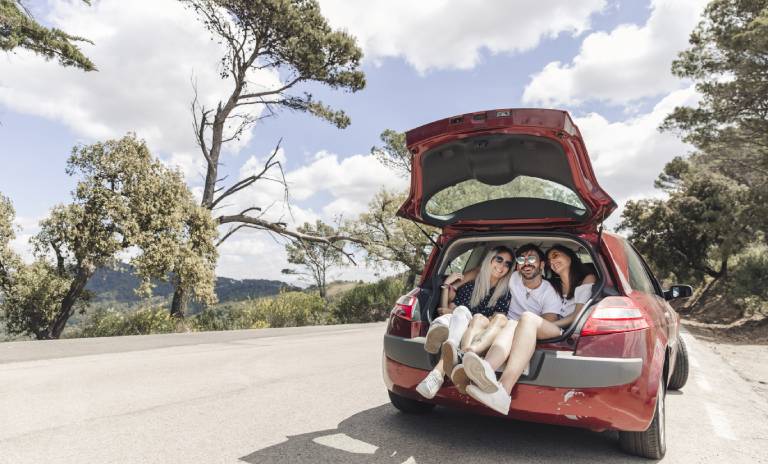 Tres personas sentadas en la parte trasera de un carro con gesto de felicidad y un paisaje lleno de árboles y montañas en el fondo