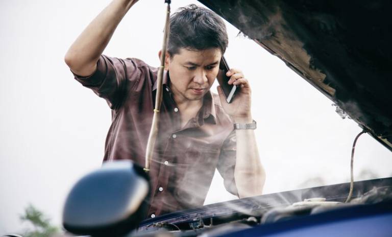 Hombre hablando por celular al lado del capó de su carro que emite humo. 
